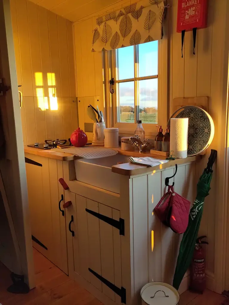 A rustic sink and food prep area bathed in warm sunlight at Eastleach Downs Organic Farm Shepherd's Hut, Gloucestershire